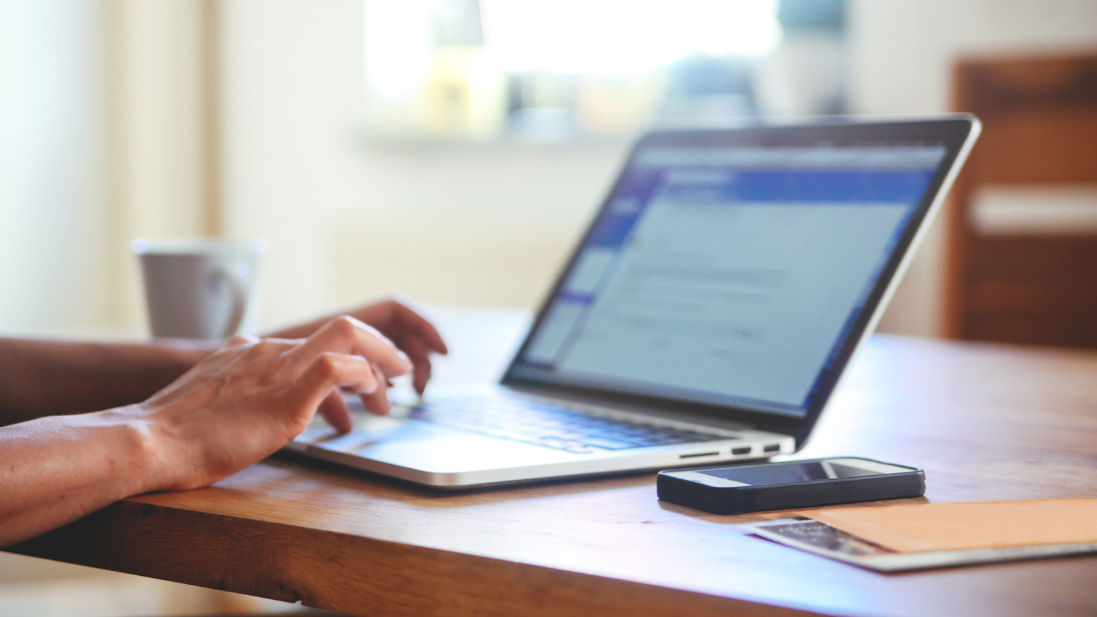 Person at table using a laptop with a smart phone, papers and coffee cup nearby.