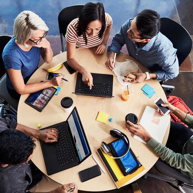 Group of employees seated at round table with laptops, tablets and smart phones brainstorming and taking notes.